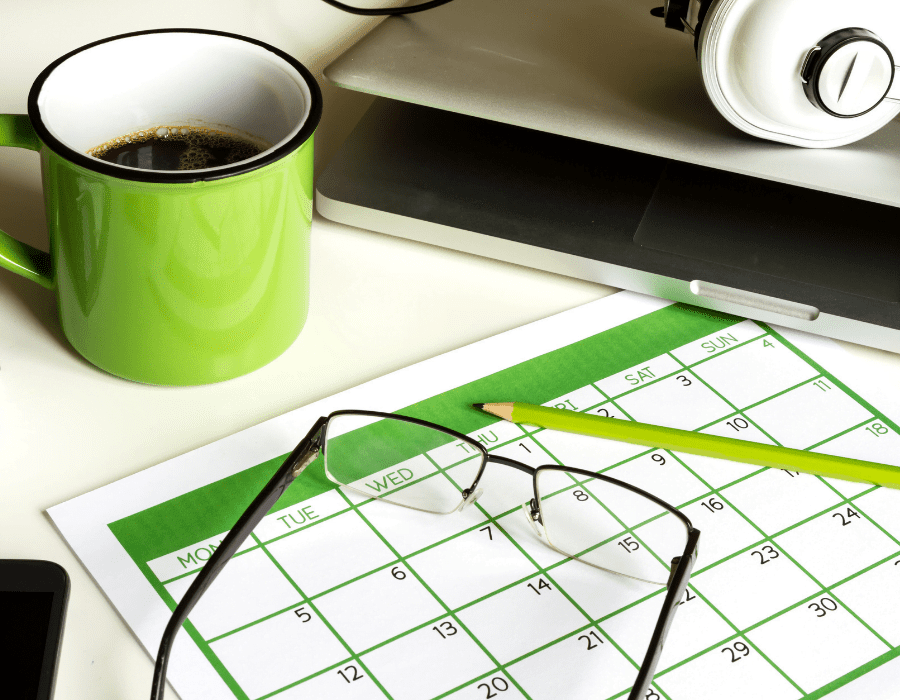 A calendar sits next to a laptop with headphones, a cup of coffee, and glasses and a pencil. Setting reasonable turnaround expectations with your virtual assistant is crucial to your working relationship and productivity.