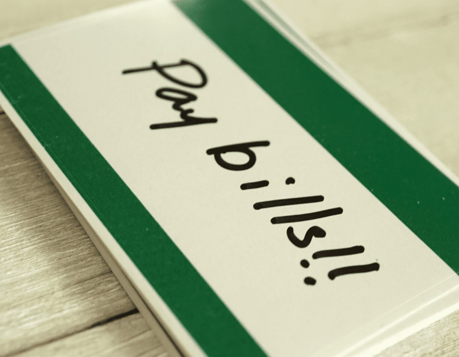 A green and white notecard features handwriting that says “pay bills!” Automating business tasks like invoicing and paying bills will help you save money and make/receive payments on time.