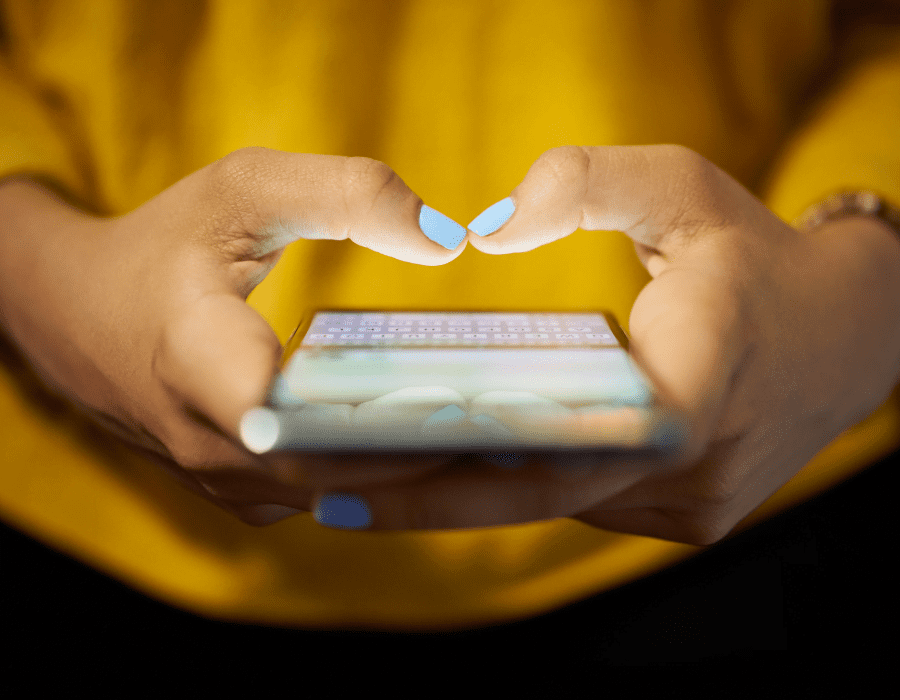 A woman in a yellow shirt holds her phone and thumbs the keyboard posting on social media. Automating social media tasks will free up a lot of time in your business.