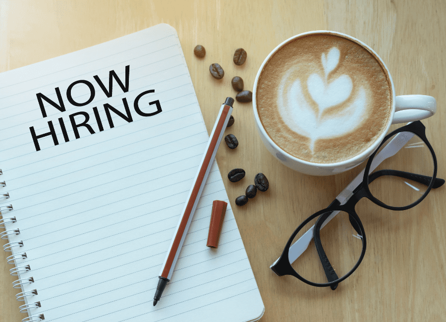 "Now Hiring" is printed on a notebook next to a mug of coffee and glasses. Erika Macauley can help you learn which team members to hire and when.